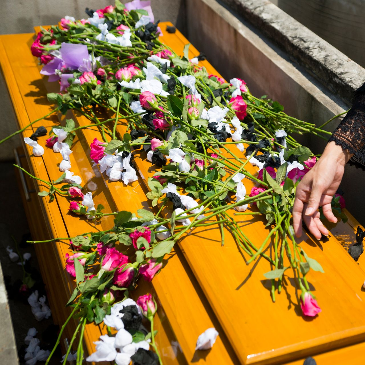 Une personne dépose des fleurs sur un cercueil en bois à l'extérieur. Des fleurs colorées recouvrent le dessus.