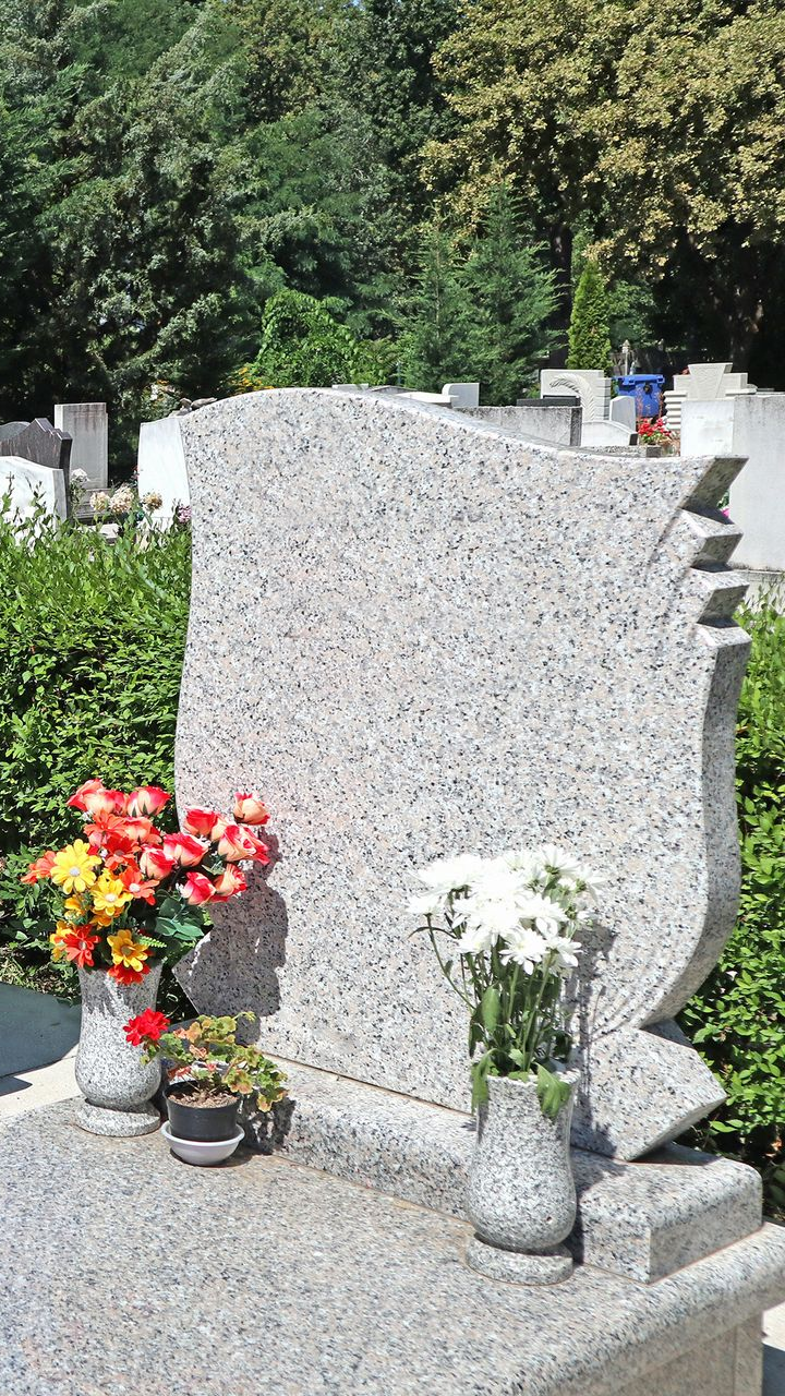 Pierre tombale fleurie dans un cimetière. Granit gris au sommet ondulé, avec des fleurs colorées à la base.