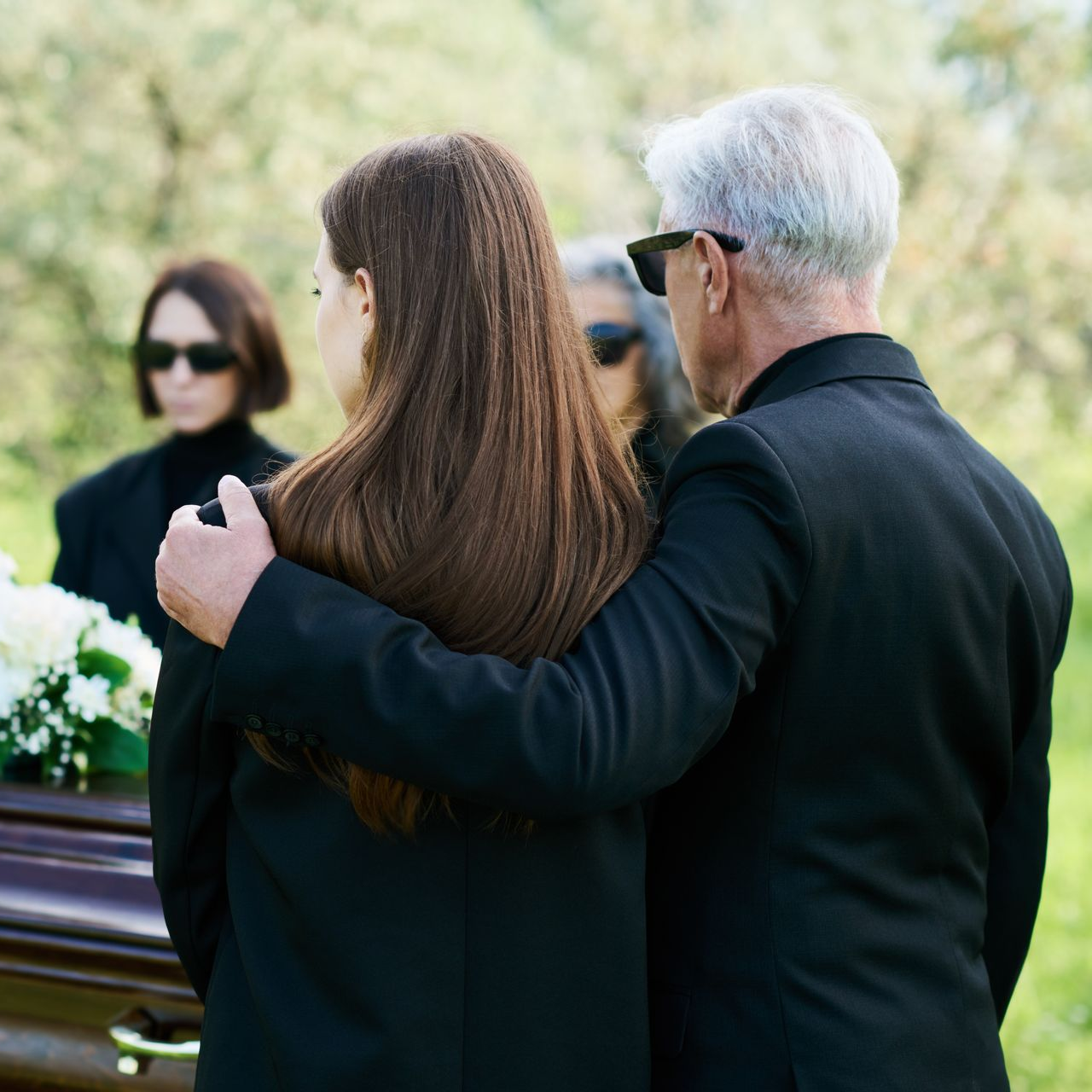 Personnes en deuil à un enterrement. Un homme réconforte une femme près du cercueil, d'autres personnes en deuil se tiennent à proximité. Décor extérieur.