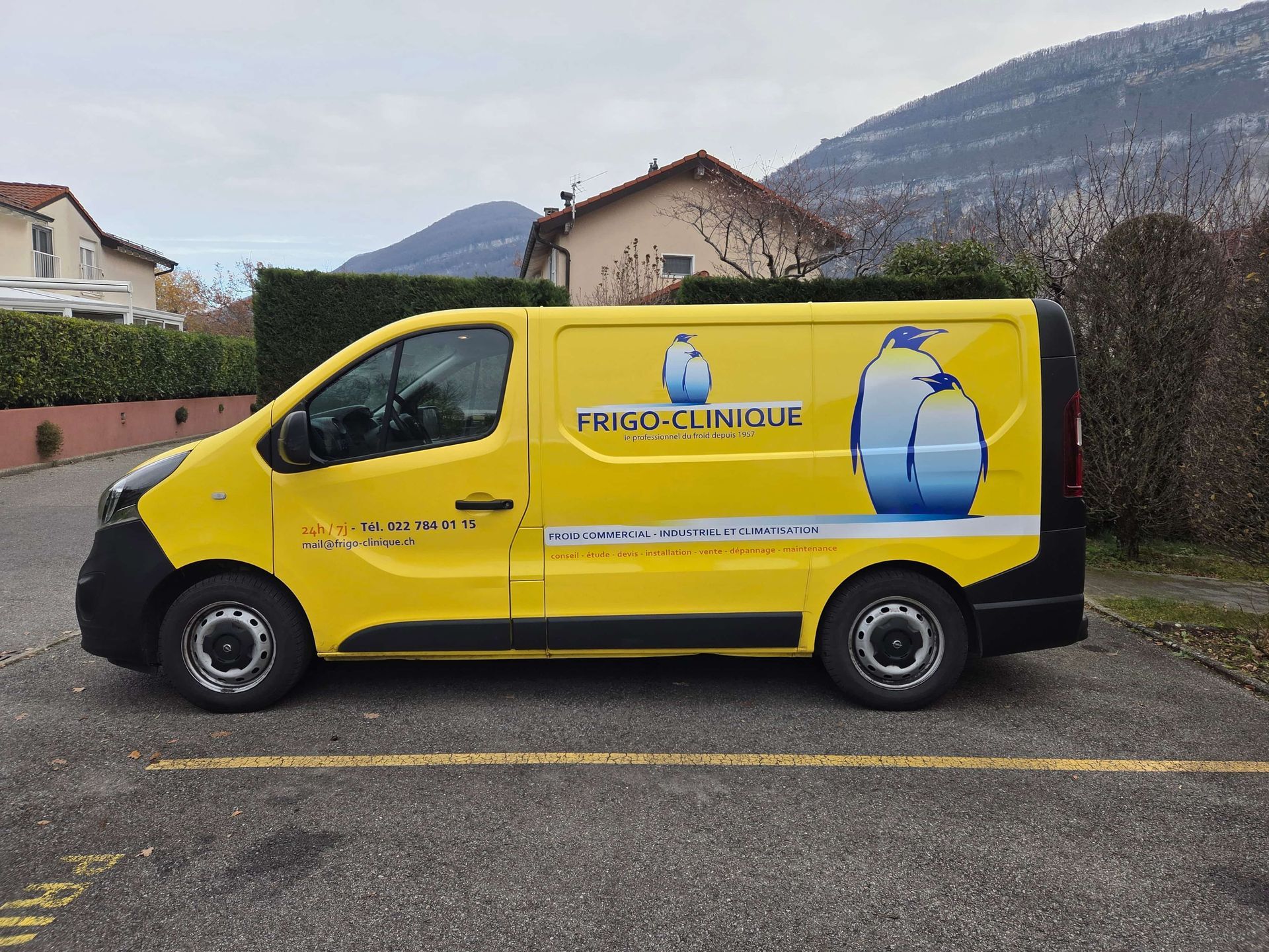 Une camionnette de livraison jaune ornée d'un logo de bouteille d'eau est garée dans une rue, devant des maisons et des montagnes.