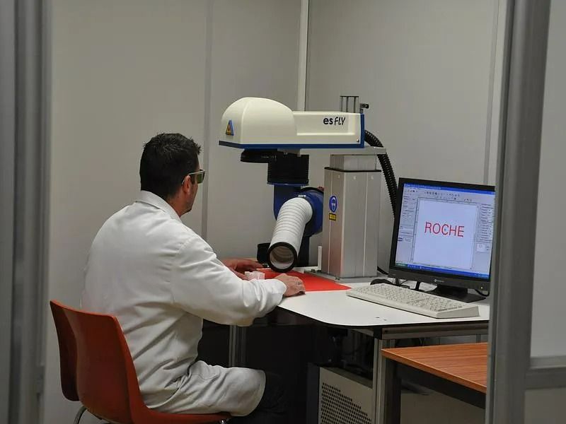 Technicien en blouse blanche utilisant un bras robotisé à un poste de travail équipé d'un écran d'ordinateur.