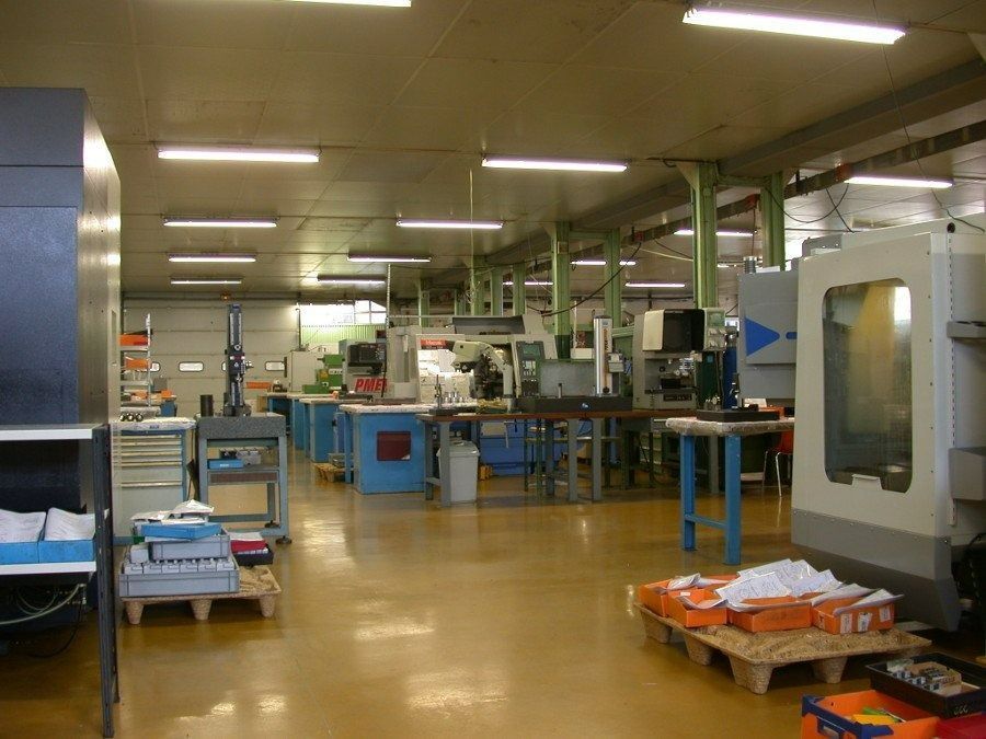 Atelier industriel avec machines, établis et piles de matériaux sous une lumière vive zénithale