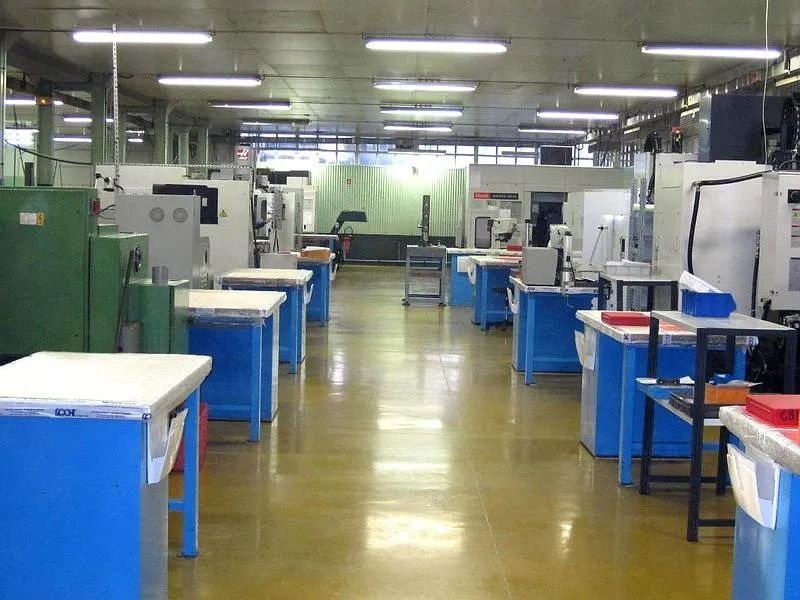 Atelier industriel lumineux avec des établis bleus et des machines disposées en rangées sous des éclairages fluorescents.