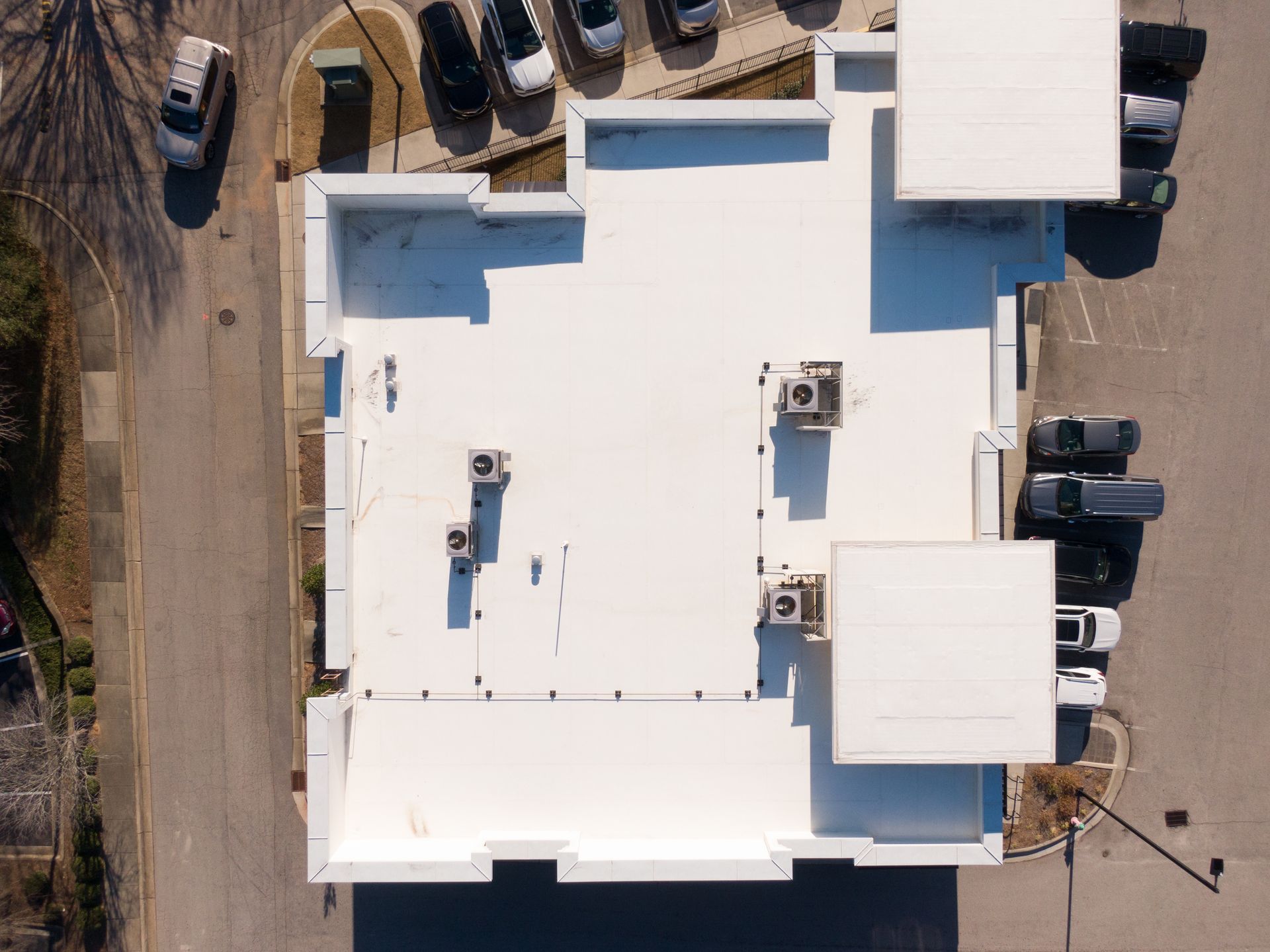Vue aérienne d'un bâtiment commercial blanc avec des unités de climatisation sur le toit, entouré de parkings et de voitures.