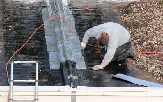 Couvreur sur un toit plat, appliquant des solins autour d'une ventilation. Présence d'un câble électrique orange et d'une échelle.