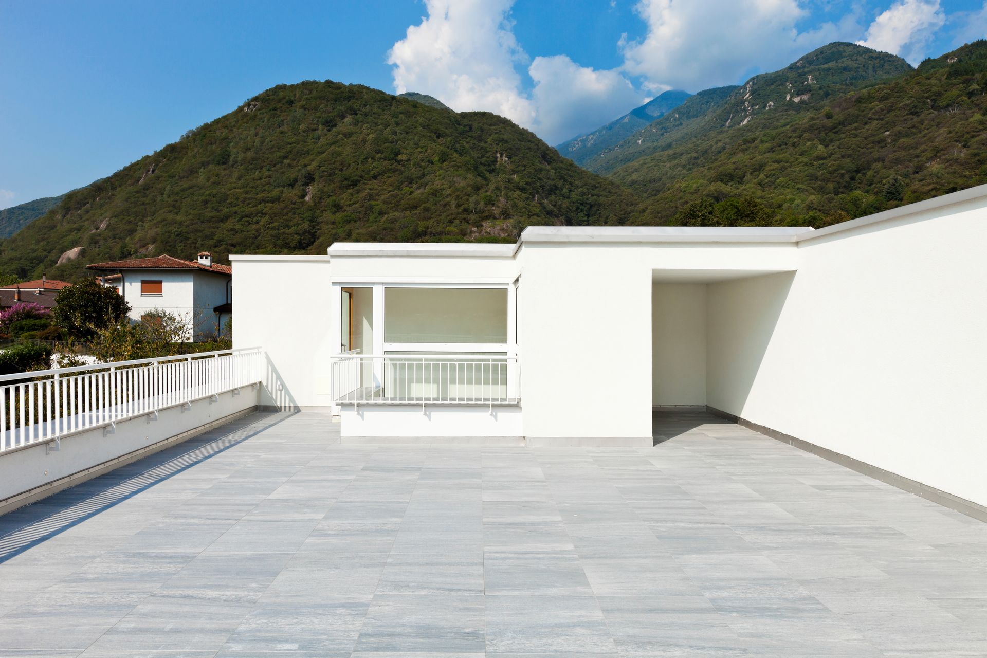 Bâtiment moderne blanc avec terrasse carrelée de gris, balustrade et vue sur les montagnes.