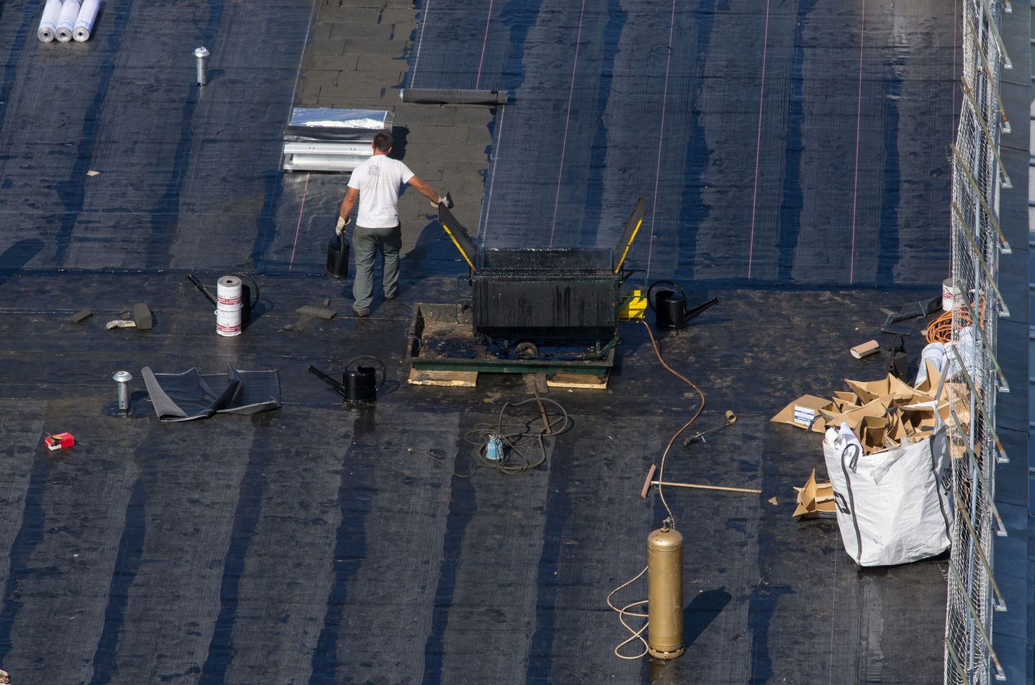 Un couvreur travaille sur un toit plat sombre et humide, près d'outils, d'une grande cuve et d'un sac blanc, avec un échafaudage à droite.