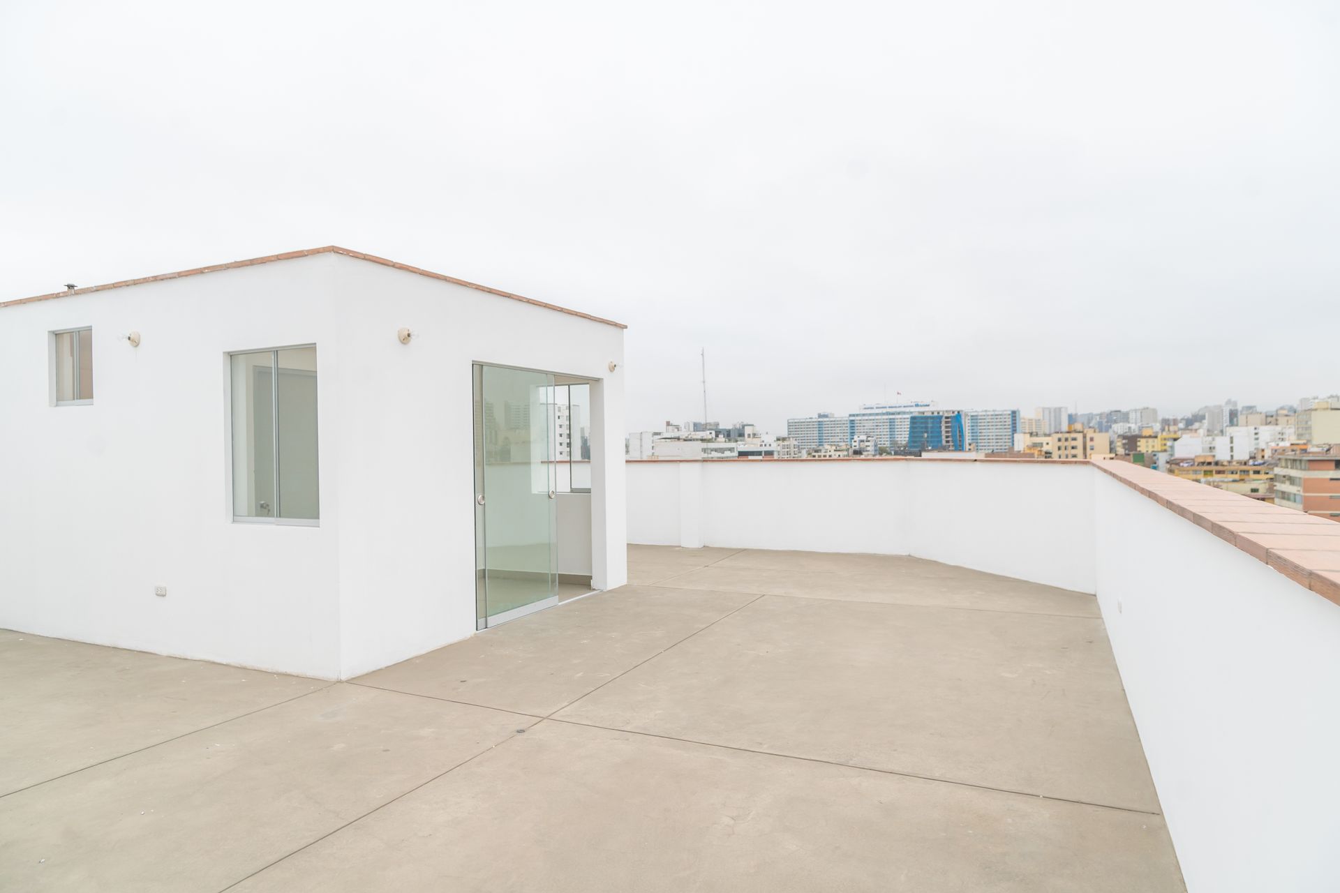 Toit blanc avec un petit bâtiment, portes vitrées et vue sur la ville sous un ciel couvert.