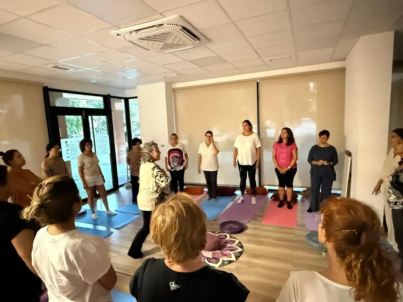 Personas de pie en círculo sobre esterillas de colores en un estudio de yoga, aparentemente en una clase grupal.