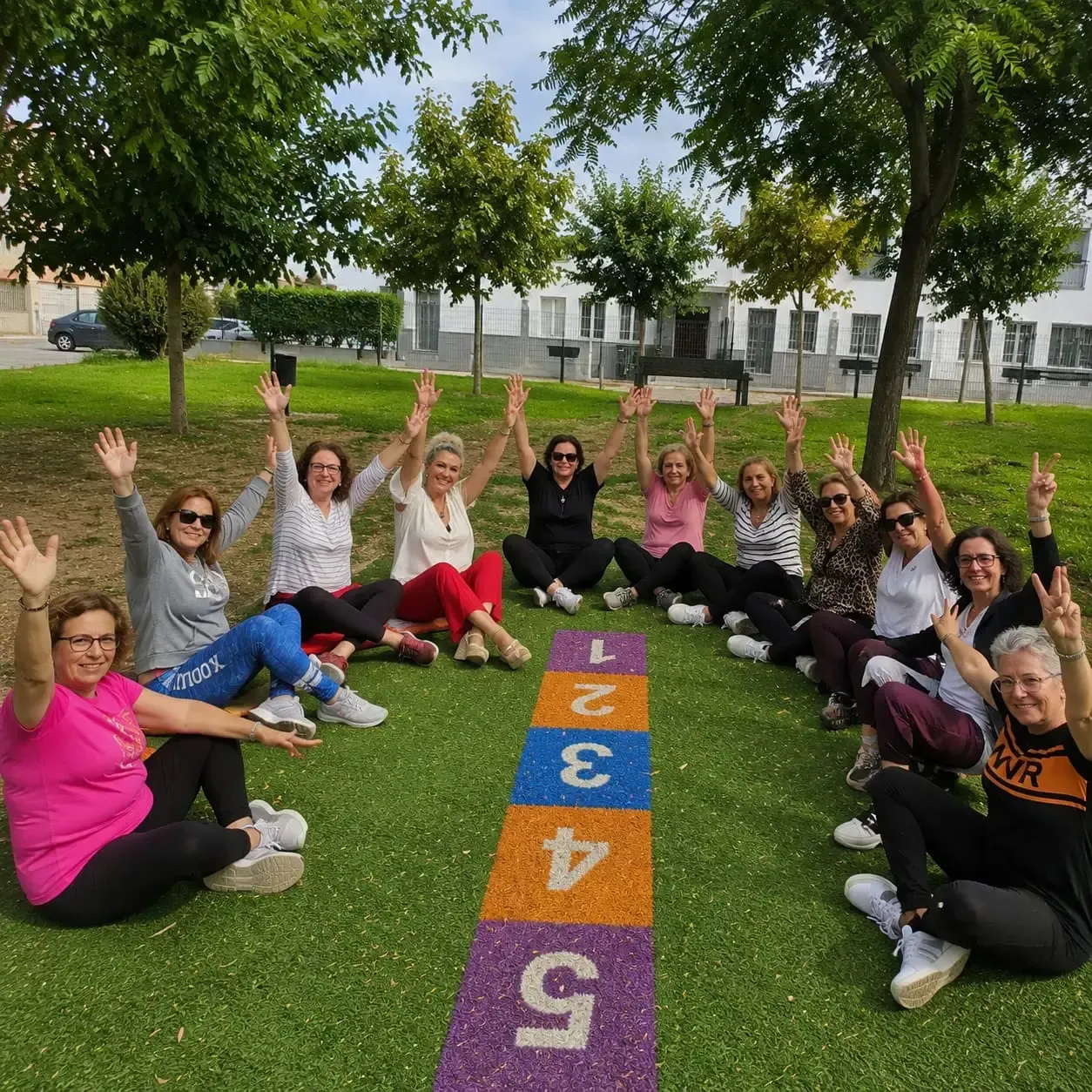 Grupo sentado en un parque, sonriendo y saludando alrededor de un colorido circuito de rayuela numerado.