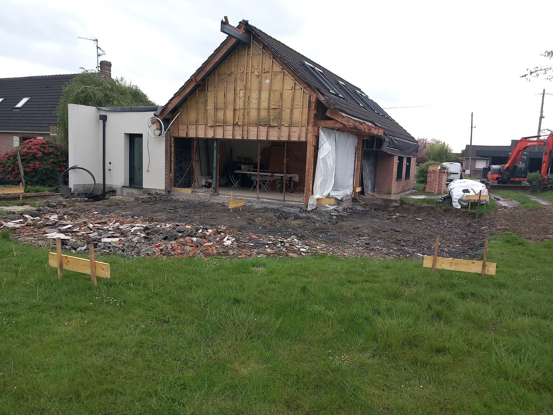 Bâtiment en briques partiellement démoli avec intérieur exposé, entouré de débris, dans une cour herbeuse.
