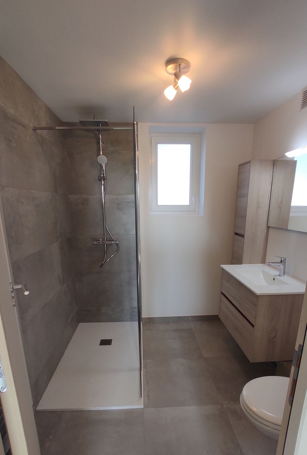 Salle de bains avec douche, lavabo et fenêtre ; la douche comprend un siège rabattable et une barre d'appui