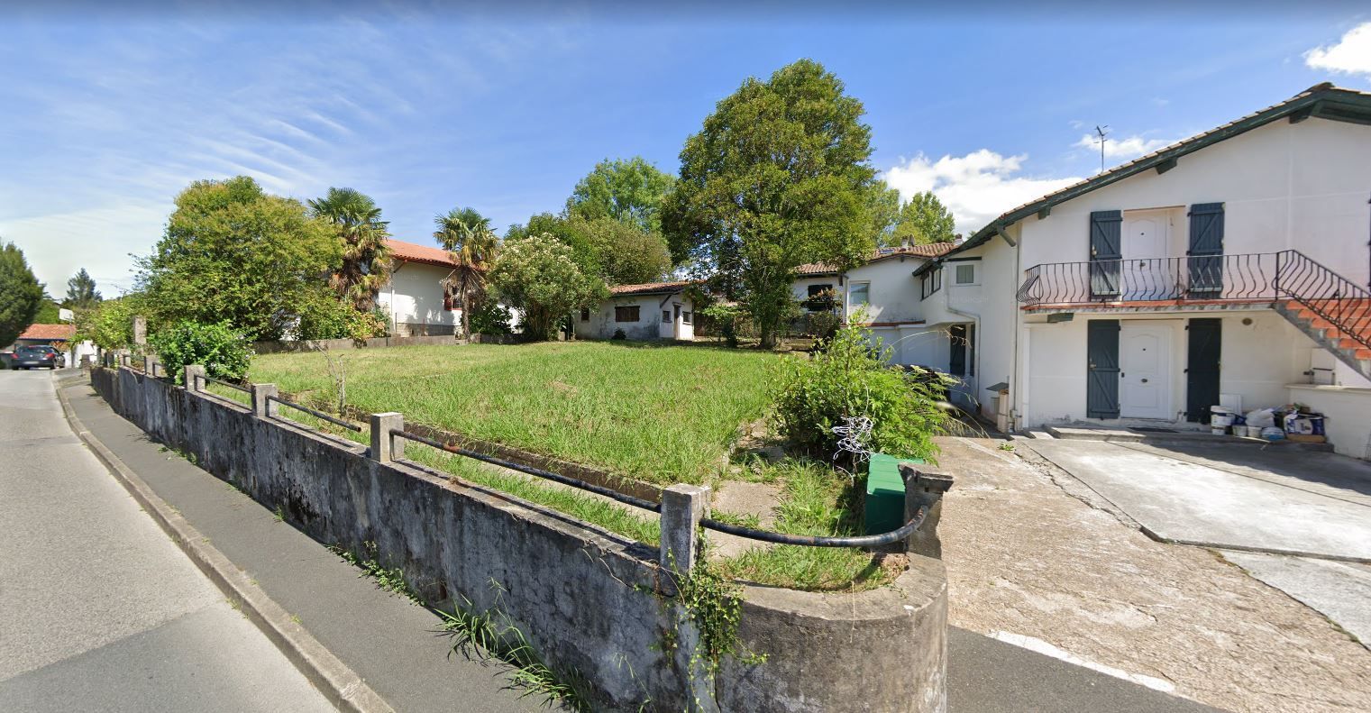 Rue résidentielle ensoleillée avec une pelouse, un mur de pierre, des arbres et des maisons blanches avec balcons.