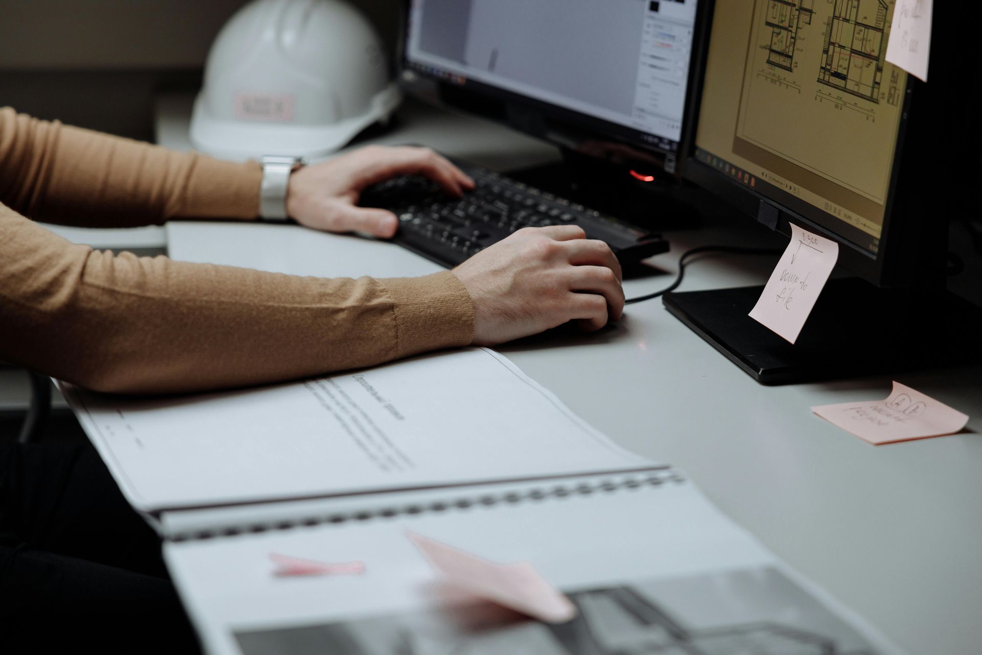 Des mains tapant sur un clavier à un bureau, un cahier ouvert, un écran d'ordinateur, un clavier et des notes adhésives roses.