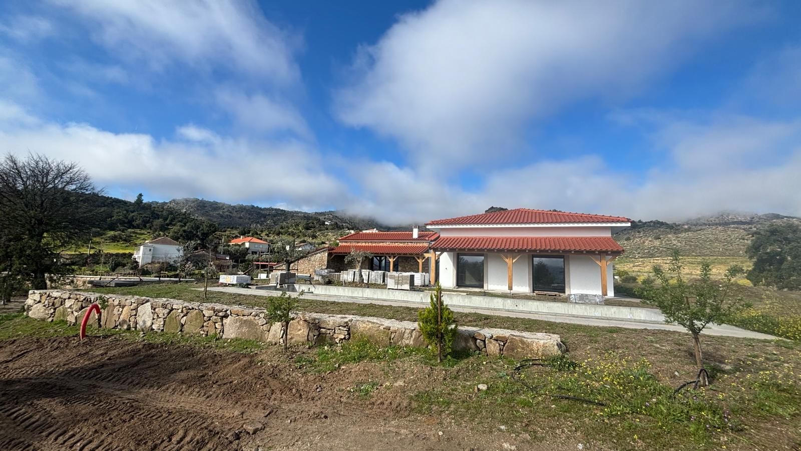 Une maison au toit rouge et aux colonnes blanches se dresse dans un paysage rural sous un ciel nuageux.