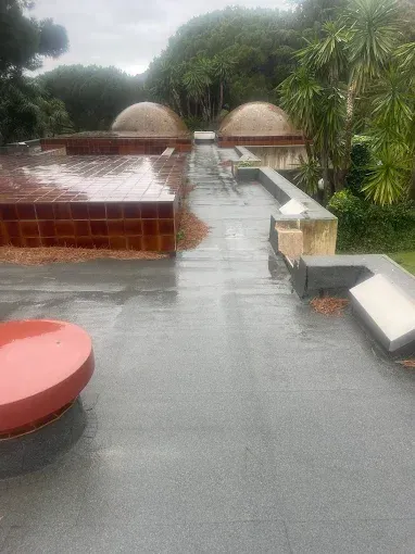 Terraza en la azotea bajo la lluvia, con mesas rojas, tumbonas blancas y frondosos árboles verdes al fondo.