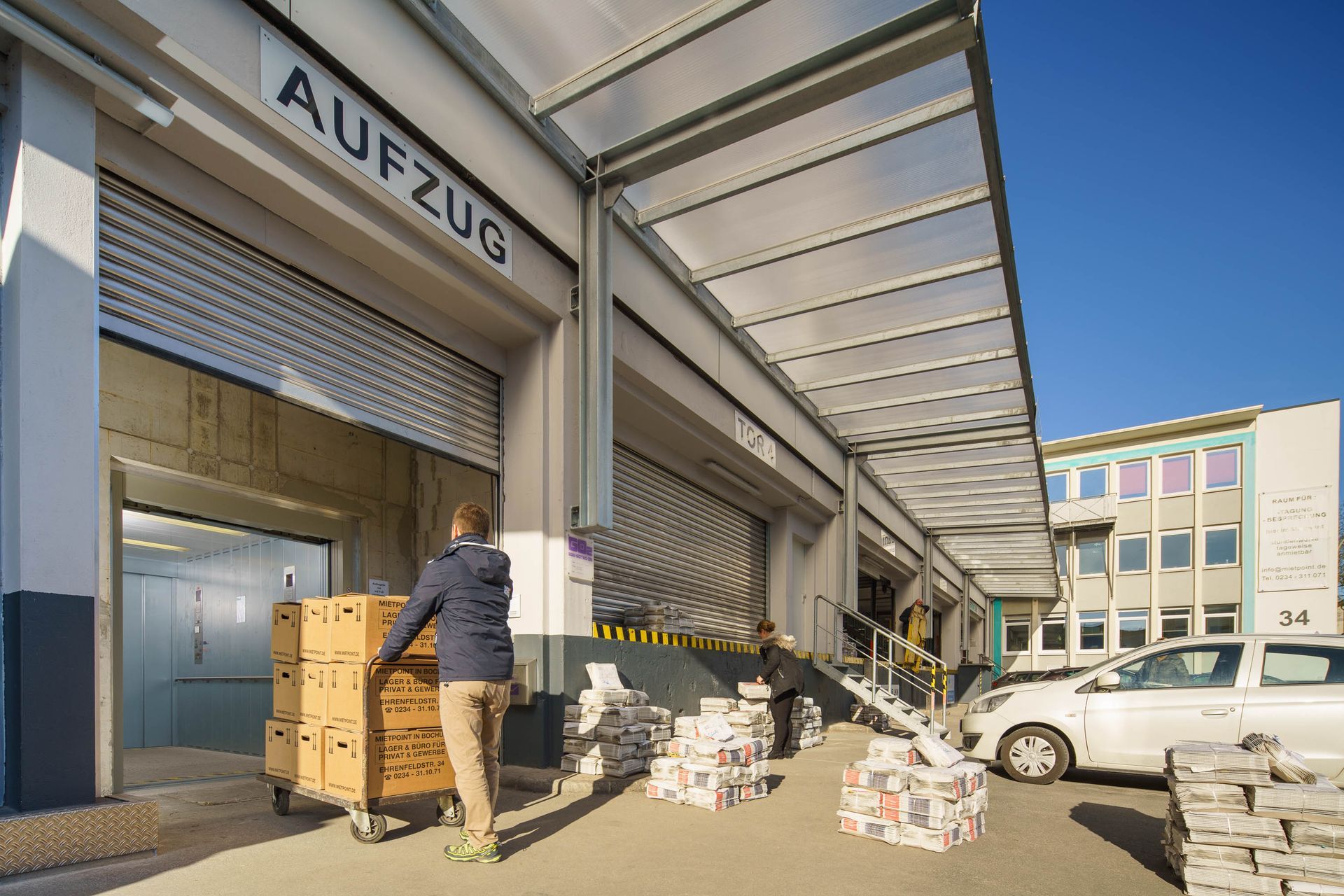 Ein Mann schiebt Kisten auf einem Rollwagen an einer Laderampe, über deren Tür das Wort „AUFZUG“ steht.