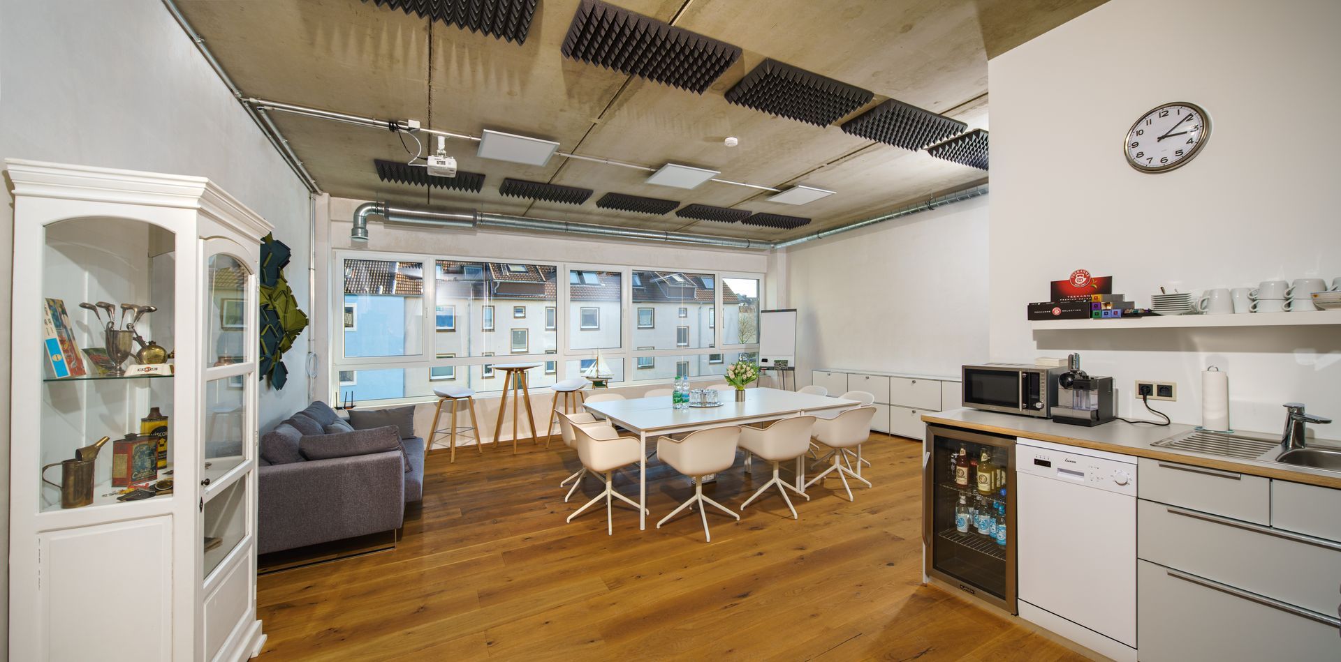 Ein lichtdurchfluteter Büroraum mit Esstisch, Stühlen, Küche und Vitrine mit Gegenständen.