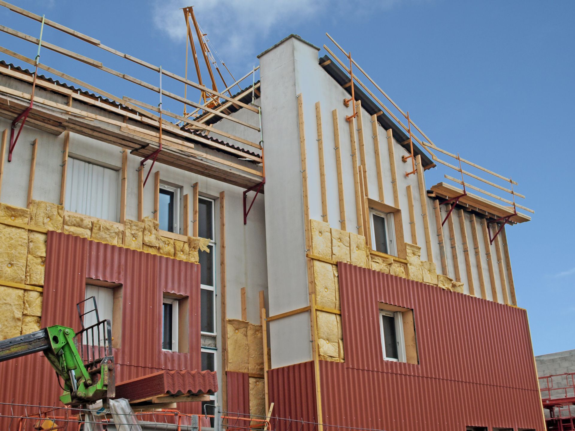 Bâtiment en construction ; mur extérieur recouvert de tôle ondulée rouge, isolation et échafaudages.