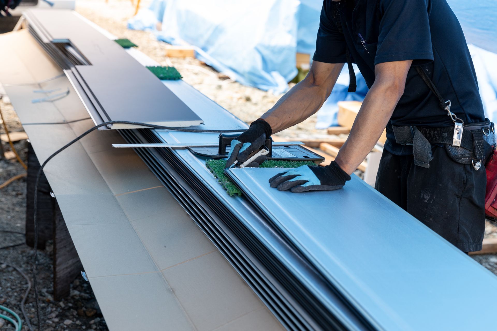 Un ouvrier portant des gants découpe une longue tôle sur une table de travail à l'extérieur.