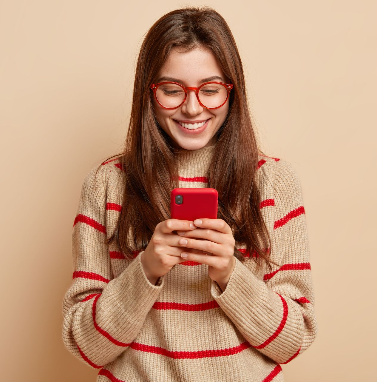 Une femme à lunettes sourit à son smartphone rouge. Elle porte un pull beige à rayures.