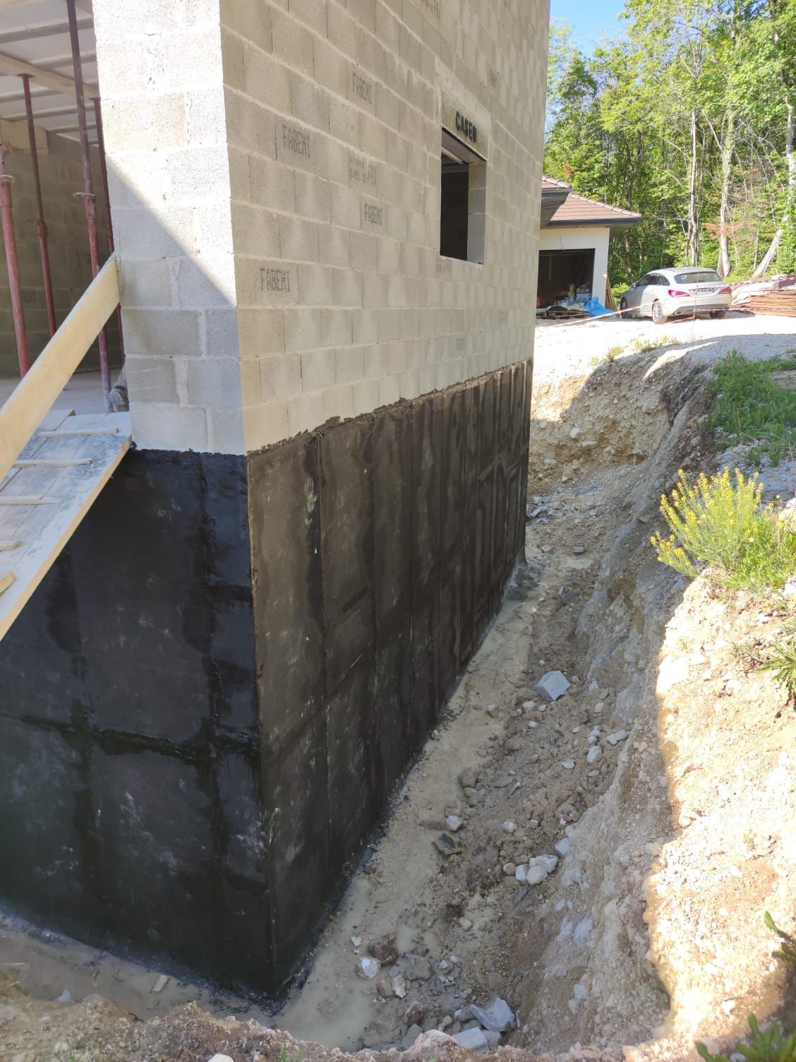 Chantier de construction avec un bâtiment en bloc de béton et une membrane d'étanchéité le long des fondations.