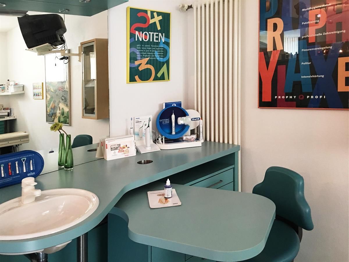Zahnarztpraxis mit Waschbecken, Arbeitsplatte, Stuhl und Geräten. Kunstwerke schmücken die Wände und das Farbschema ist blaugrün und weiß.