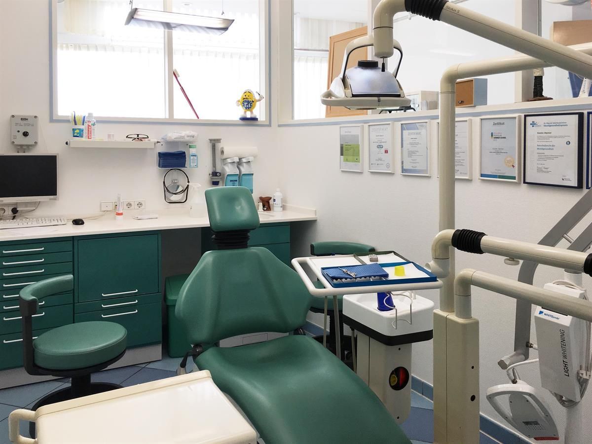 Die Inneneinrichtung einer Zahnarztpraxis mit einem grünen Zahnarztstuhl, Geräten und Schränken. Der Raum ist hell, an der Wand hängen gerahmte Zertifikate.