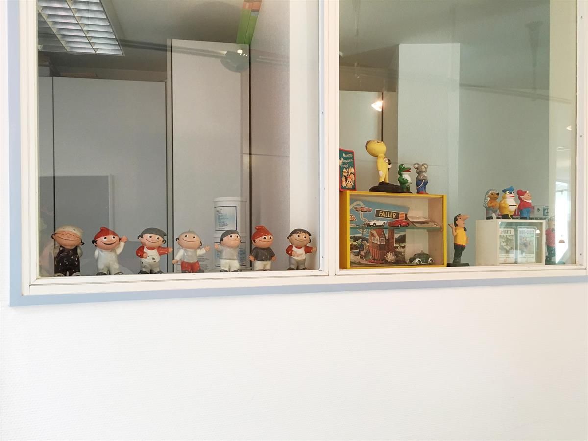 Eine Reihe kleiner Figuren in einer Vitrine mit weißem Rahmen. Weitere Gegenstände, darunter ein kleines gelbes Regal, spiegeln sich im Fenster.