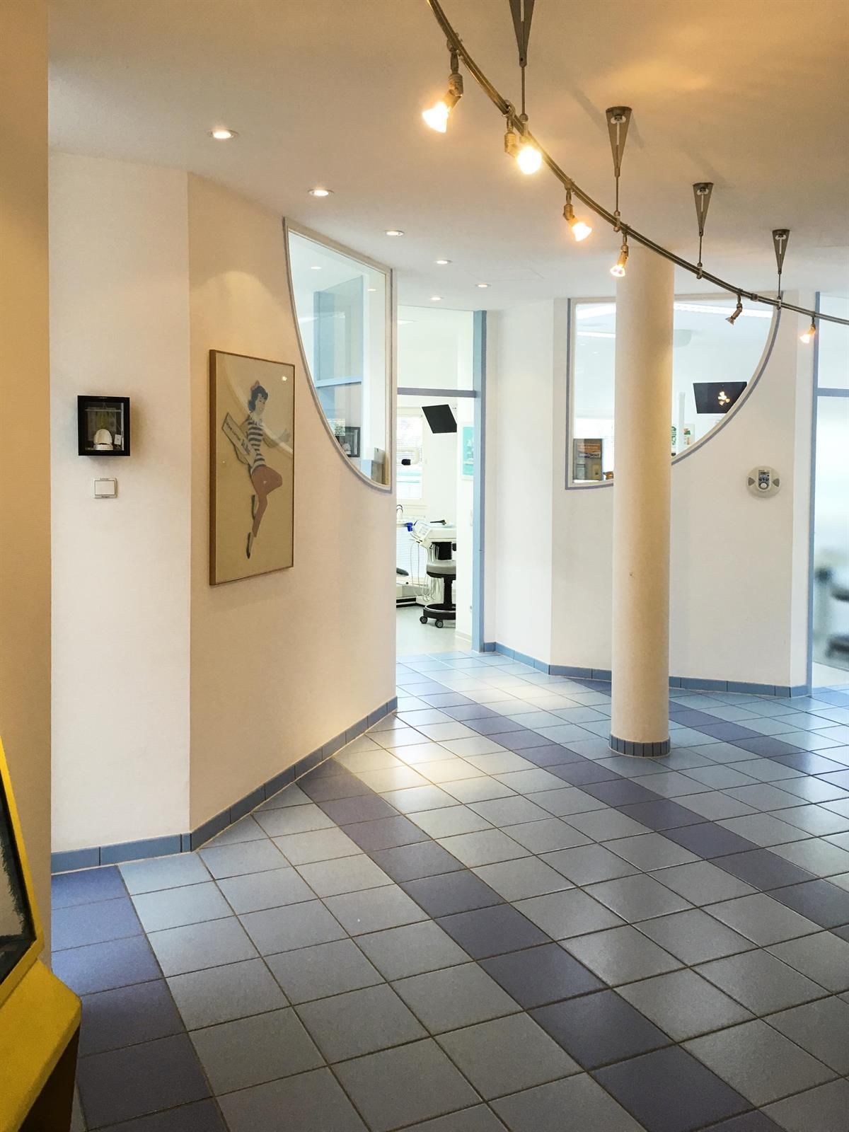 Ein Innenflur mit hellblau-weiß gefliestem Boden, weißen Wänden und Schienenbeleuchtung. Kunstwerke und geschwungene Fensterrahmen sind sichtbar.