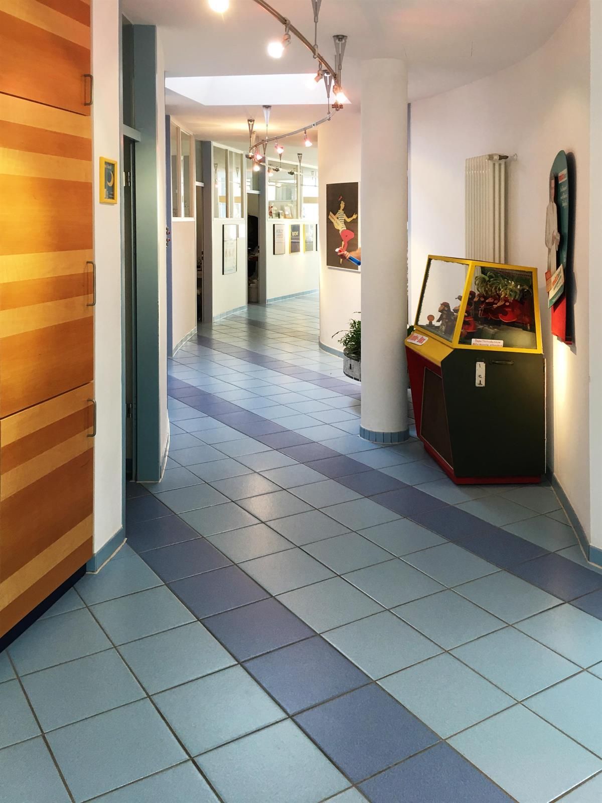 Flur mit blauem Fliesenboden, Vitrine mit bunten Gegenständen, Kunst an den Wänden und einer Holztür.