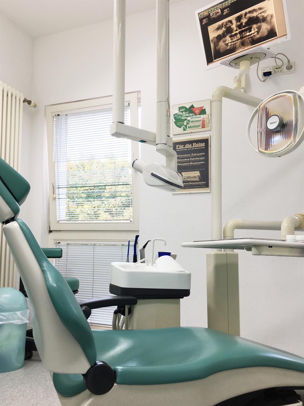 Zahnarztstuhl in einem hellen, weißwandigen Raum mit Geräten und einem Fenster. Der Stuhl ist blaugrün und der Raum wirkt sauber.