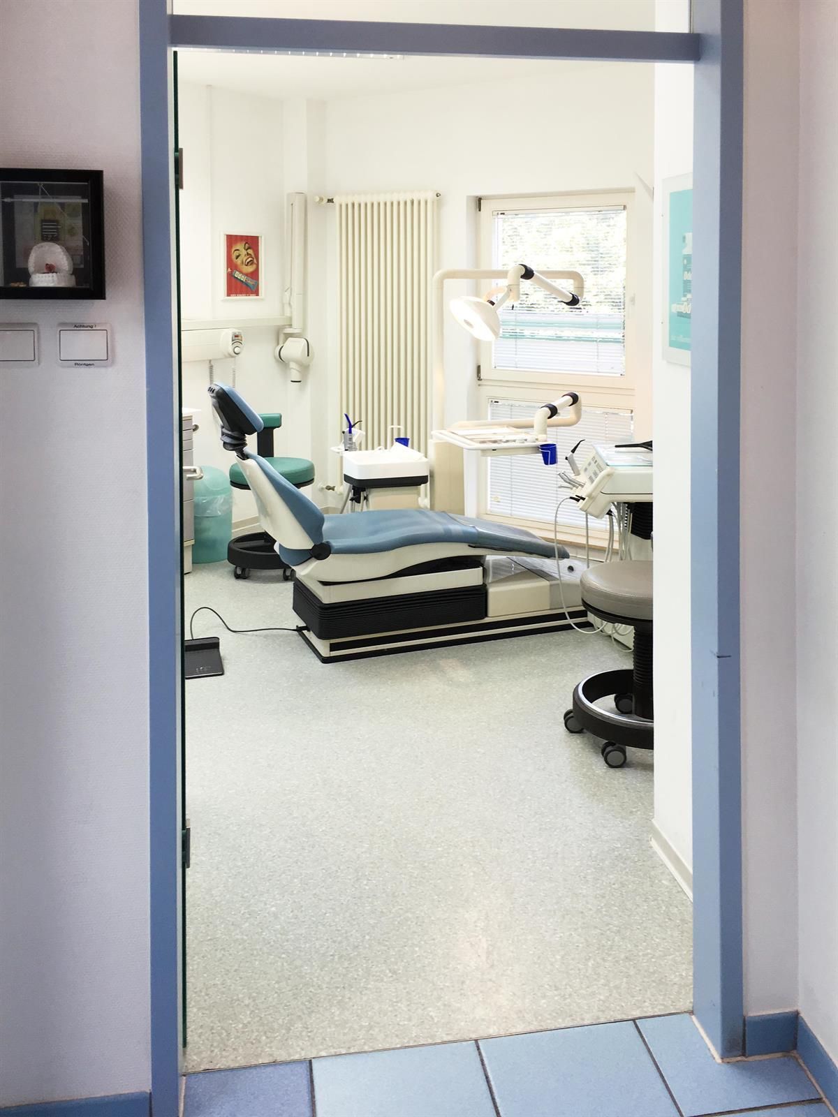 Blick in eine Zahnarztpraxis mit Zahnarztstuhl, Geräten und offener Tür. Hell erleuchteter Raum, weiße Wände und ein blau getönter Türrahmen.