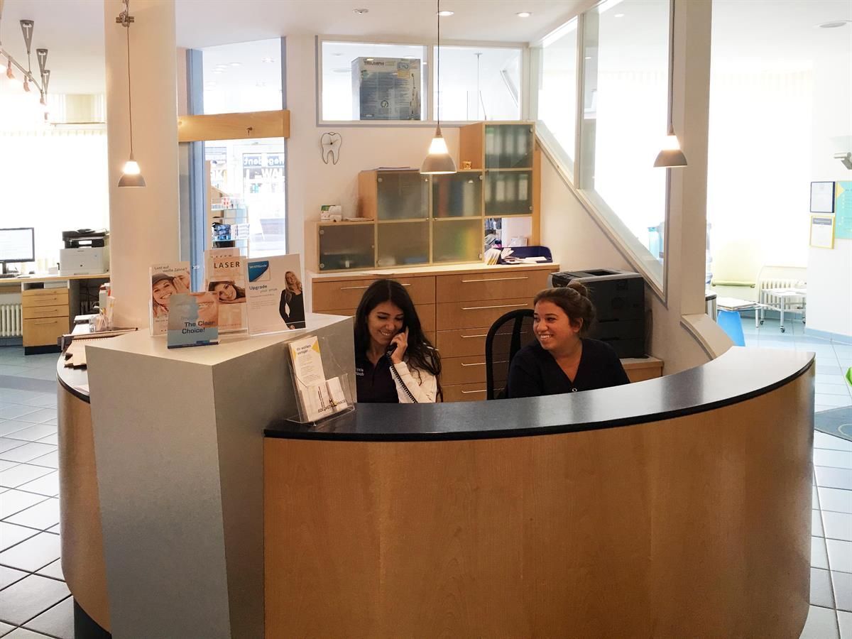 Zwei lächelnde Frauen hinter einem geschwungenen Empfangstresen in einem Büro. Eine telefoniert. Modernes, helles Büroambiente.