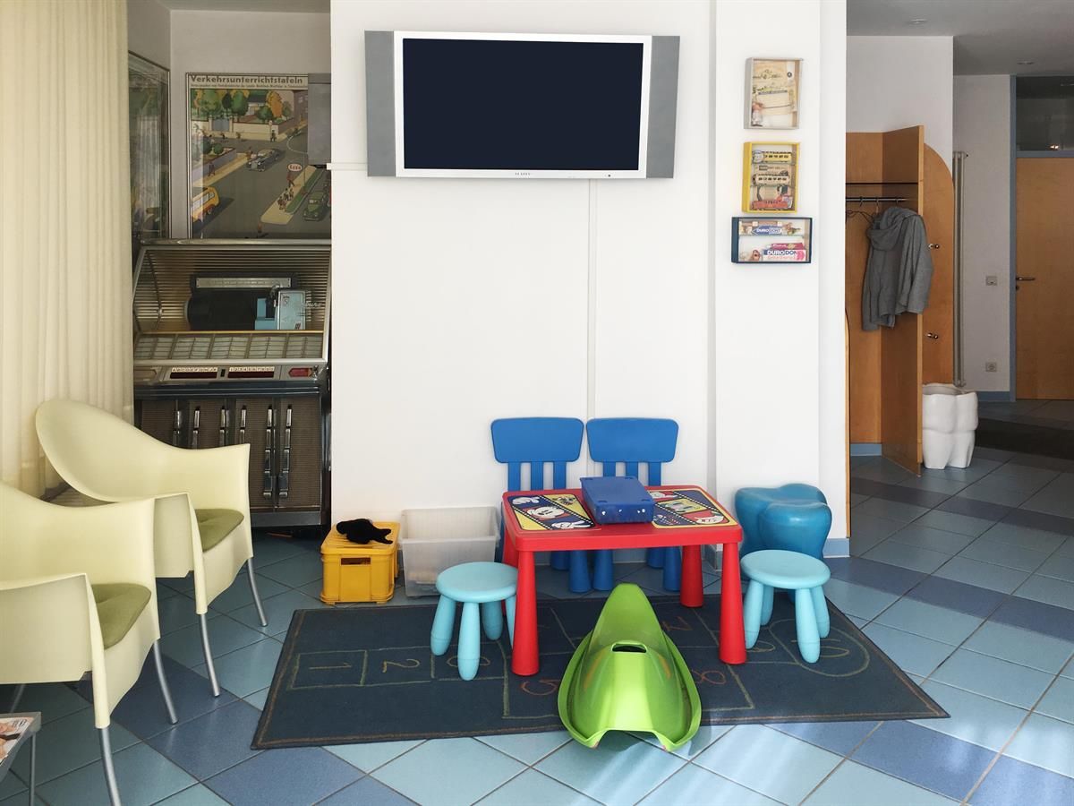 Ein kinderfreundliches Wartezimmer mit blauen und roten Möbeln, einem Fernseher und hellgrünen Stühlen.