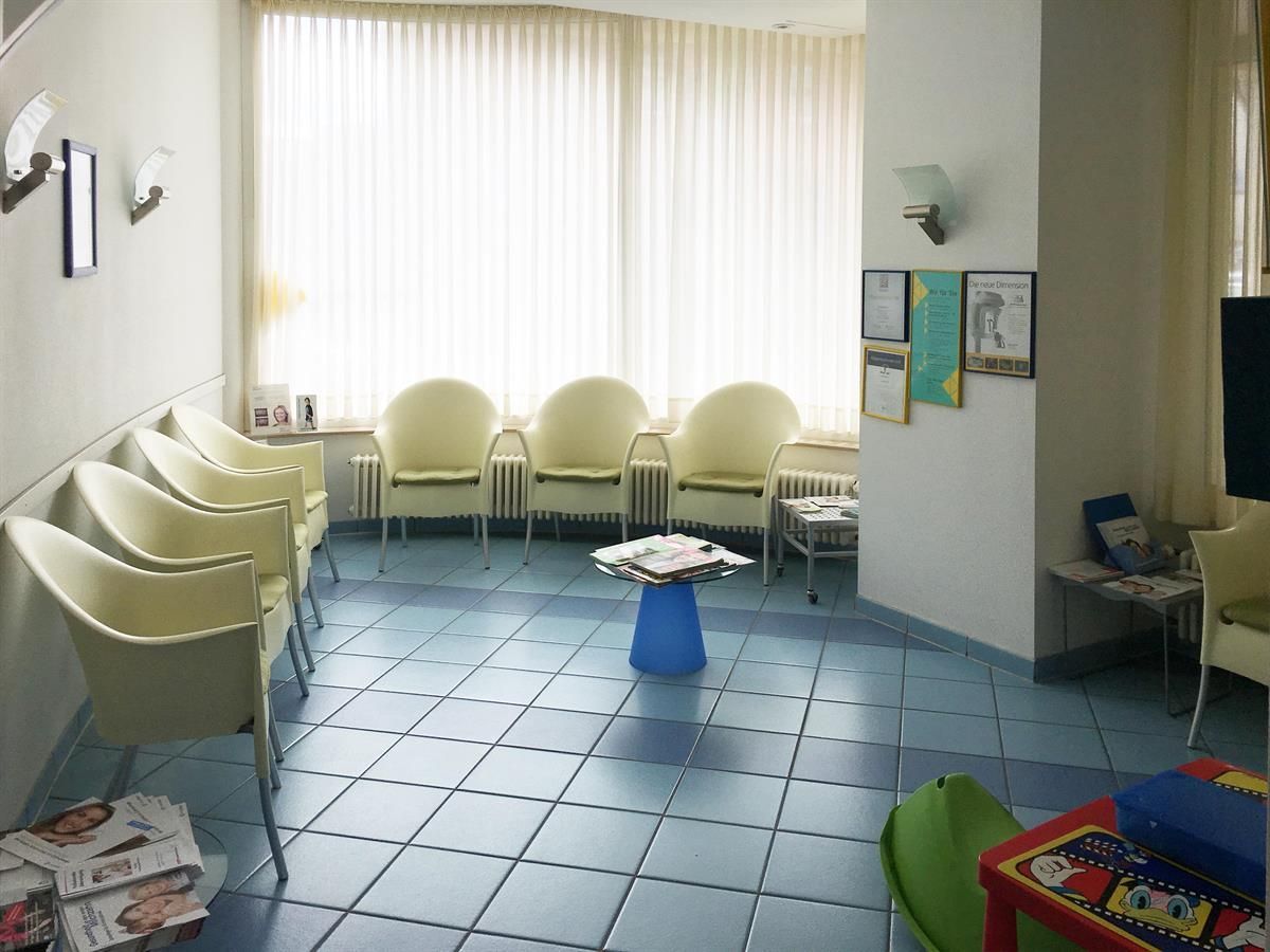 Wartezimmer mit weißen Stühlen, blauem Fliesenboden und einem Fenster mit durchsichtigen Vorhängen.