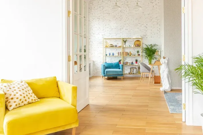Sofá amarillo, silla azul y suelo de madera en una habitación luminosa y aireada con pared de ladrillo blanco.