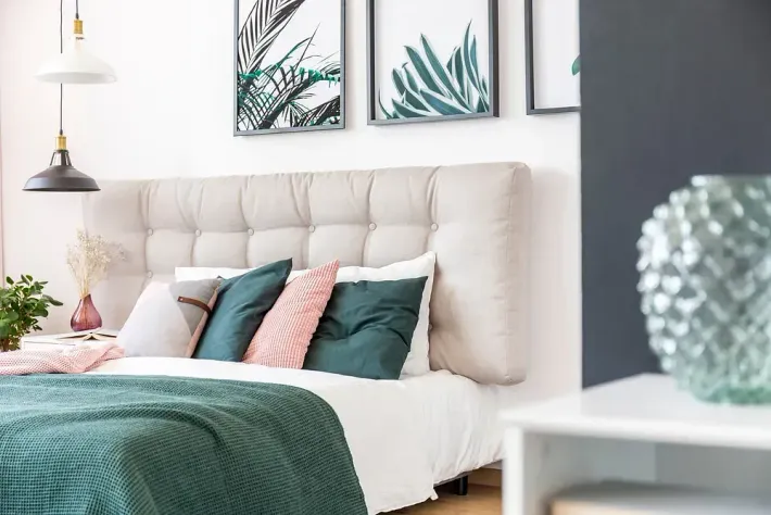 Dormitorio con cabecero tapizado, almohadas verdes y rosas y arte botánico.