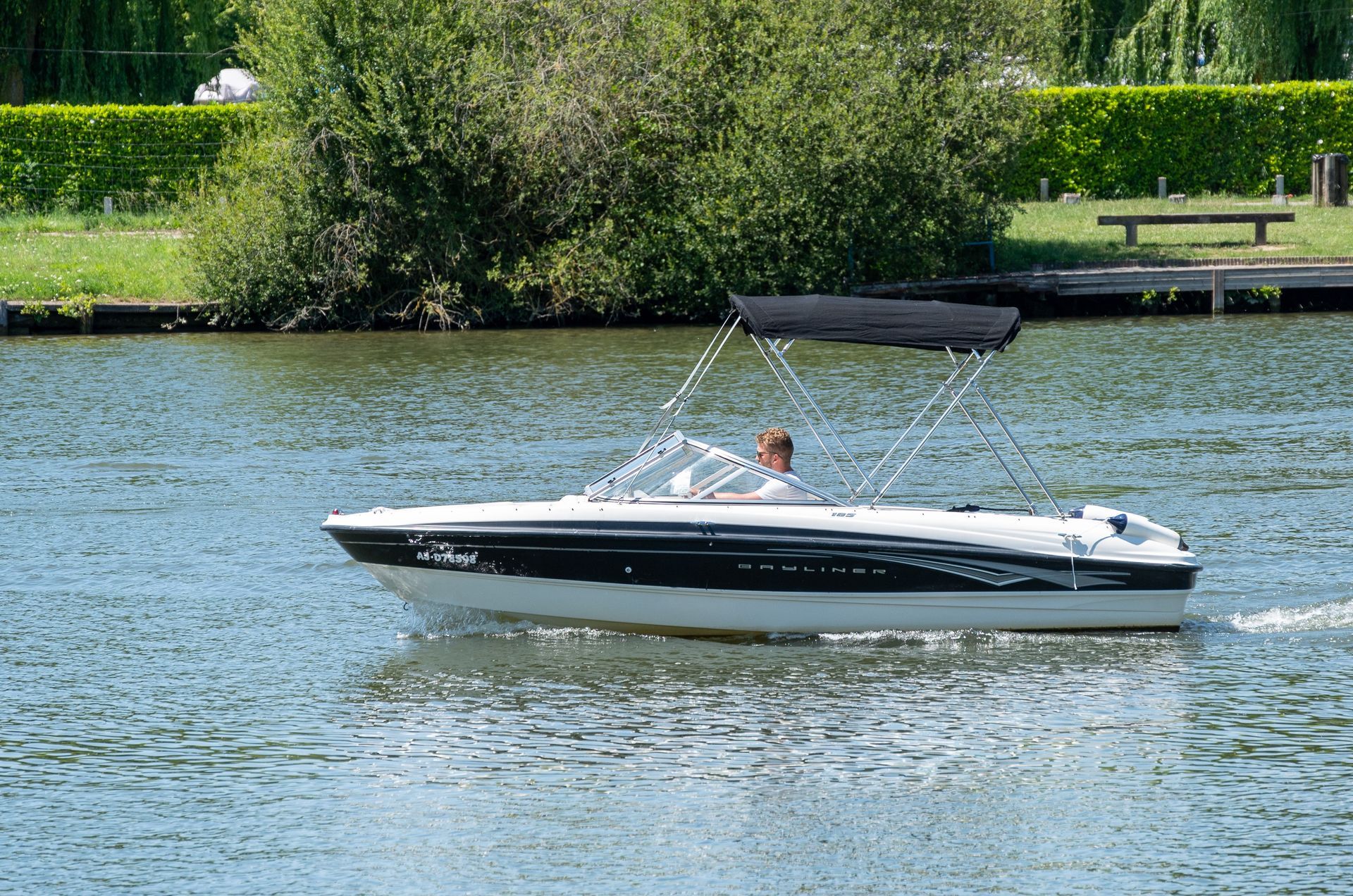 Bateau sur l'eau avec une personne au volant, coque noire et blanche, auvent, rivage vert en arrière-plan.