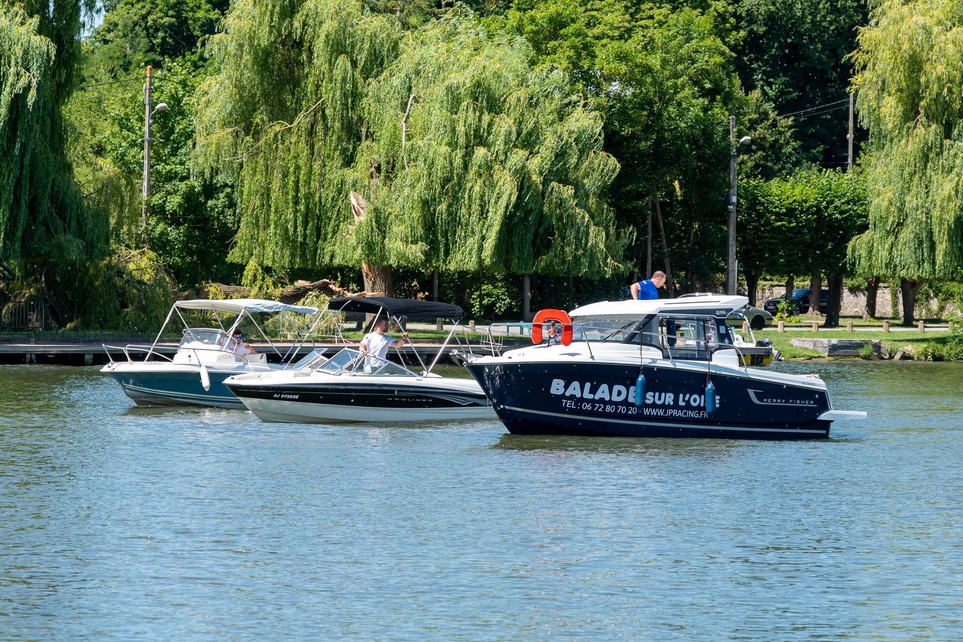 Trois bateaux à moteur sur un lac avec une rive bordée d'arbres verts par une journée ensoleillée.