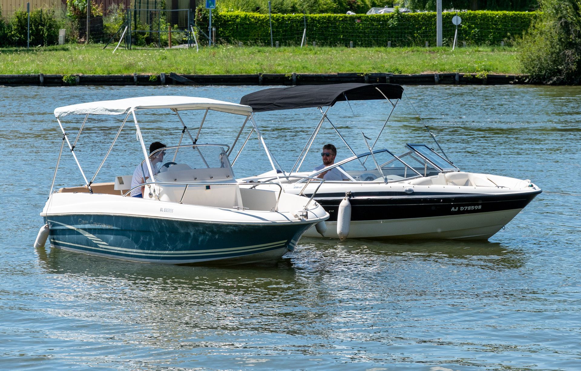 Deux bateaux à moteur sur l'eau ; l'un bleu, l'autre blanc. Des hommes pilotent les bateaux sous un ciel ensoleillé.