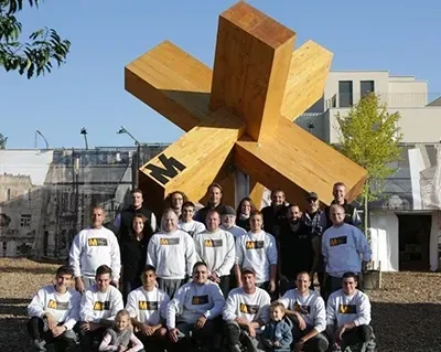 A group of people posing in front of a large wooden sculpture with the letter m on it