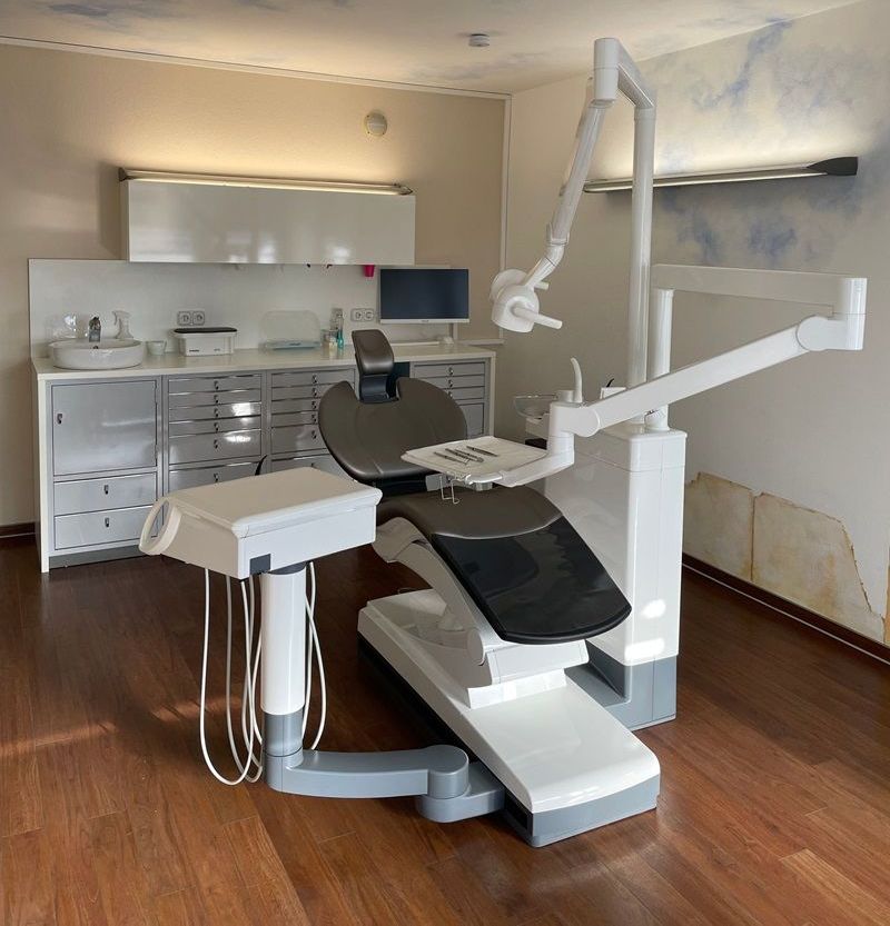 Zahnarztstuhl in einem lichtdurchfluteten Raum mit Holzboden, Schränken und einem Monitor.