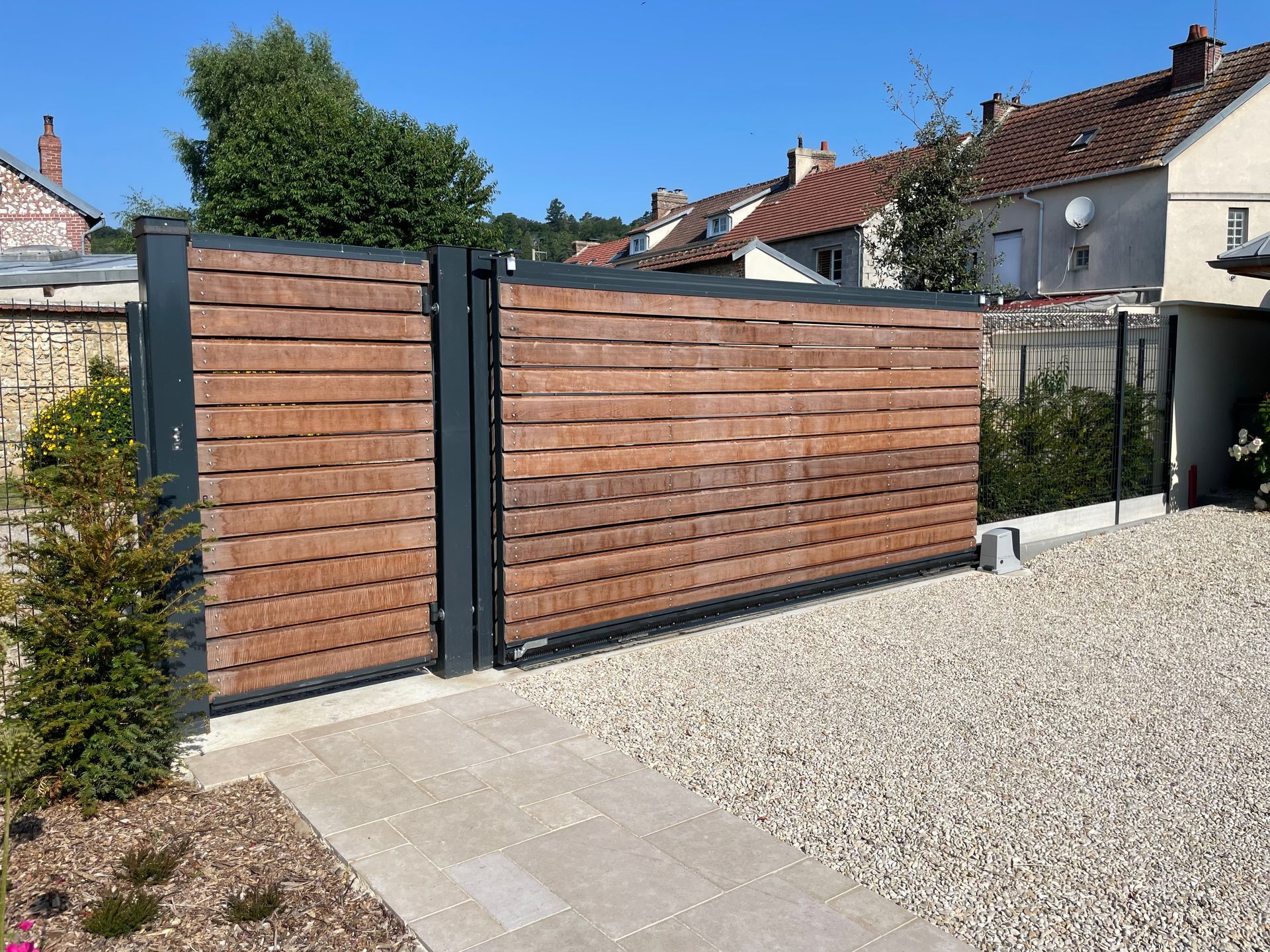 Un portail en bois à lattes, encadré d'une structure métallique grise, sur une allée de gravier devant un bâtiment.