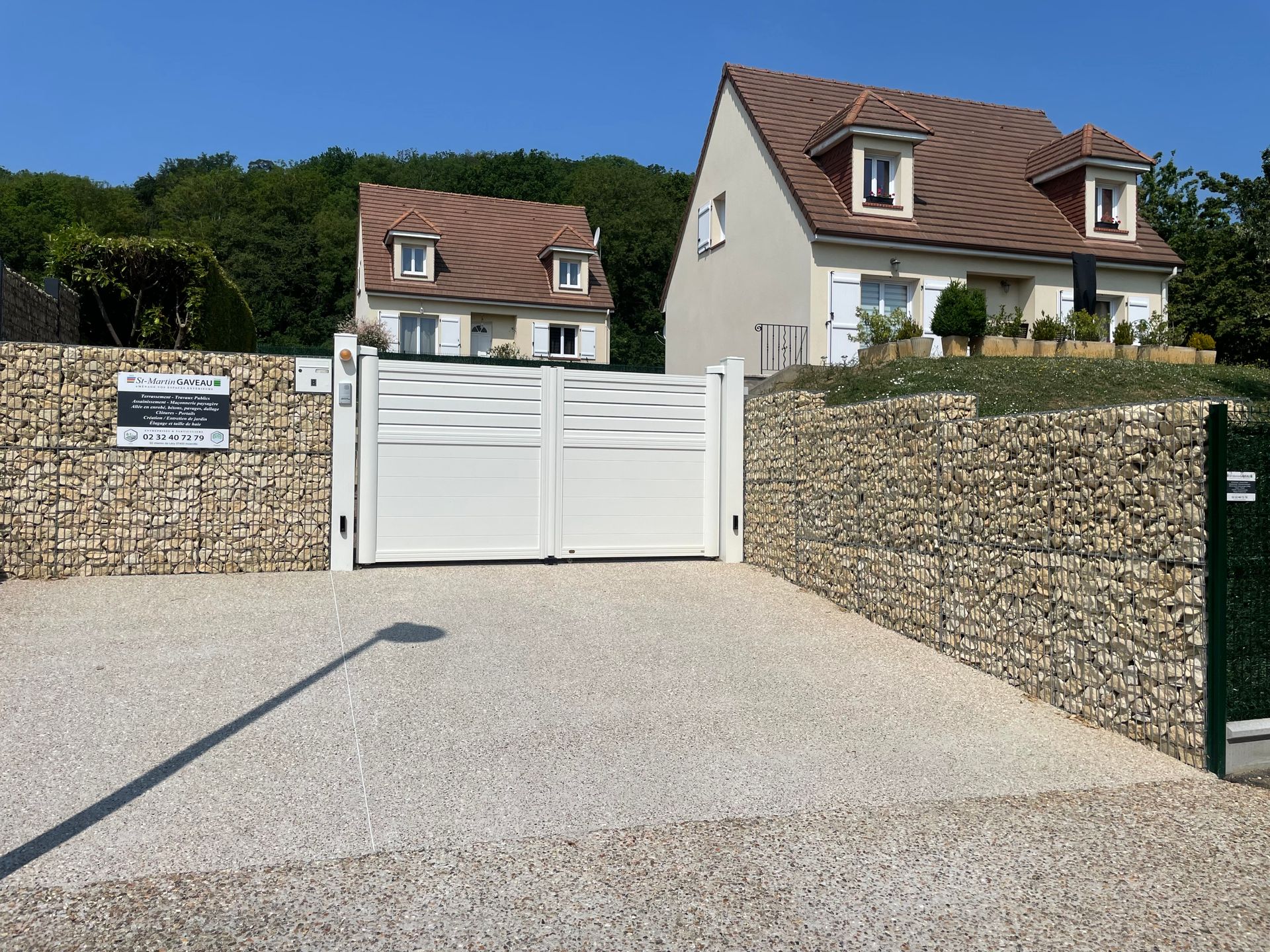 Entrée de maisons avec portail blanc, allée de gravier et murs de pierre. Deux maisons visibles à l'arrière.