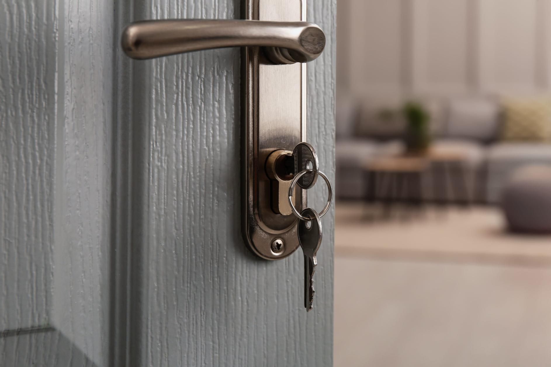 Primer plano de una puerta gris con manija de latón y llave en la cerradura, con una habitación borrosa al fondo.