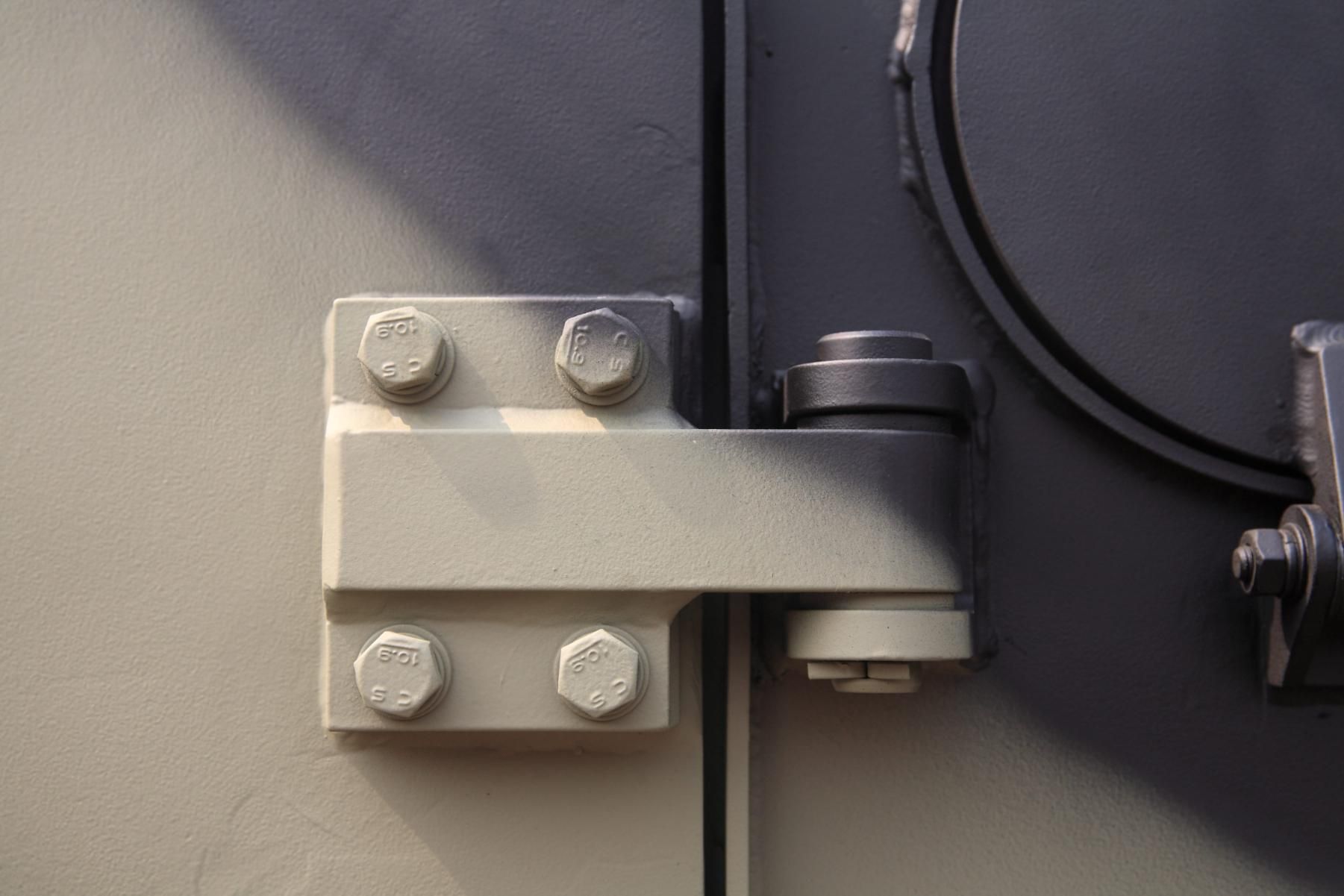 Primer plano de una bisagra o pestillo de metal gris montado en una puerta o panel de color gris oscuro.