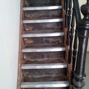 Holztreppe mit Metallstreifen auf den Stufen, dunklem Geländer und abblätternder Farbe.