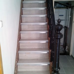 Treppe mit hellen Holzstufen, weißen Zierleisten und schwarzem Geländer.