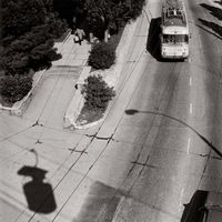 Vue aérienne d'un trolleybus sur une route, avec des lignes électriques et des ombres. Photographie en noir et blanc.